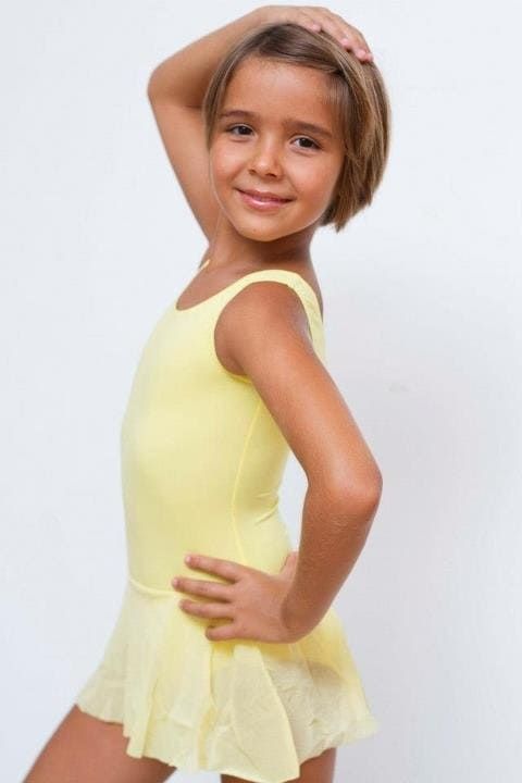 Niña con leotardo y falda de color amarillo pálido posando con una mano en la cadera sobre un fondo blanco.