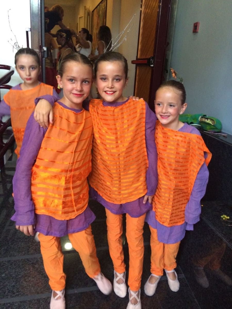 Cuatro niños disfrazados de naranja y morado posan en el interior, sonriendo y de pie juntos.