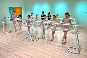 Clase de ballet practicando en una barra blanca en un estudio luminoso con paredes verde menta y suelo de madera.