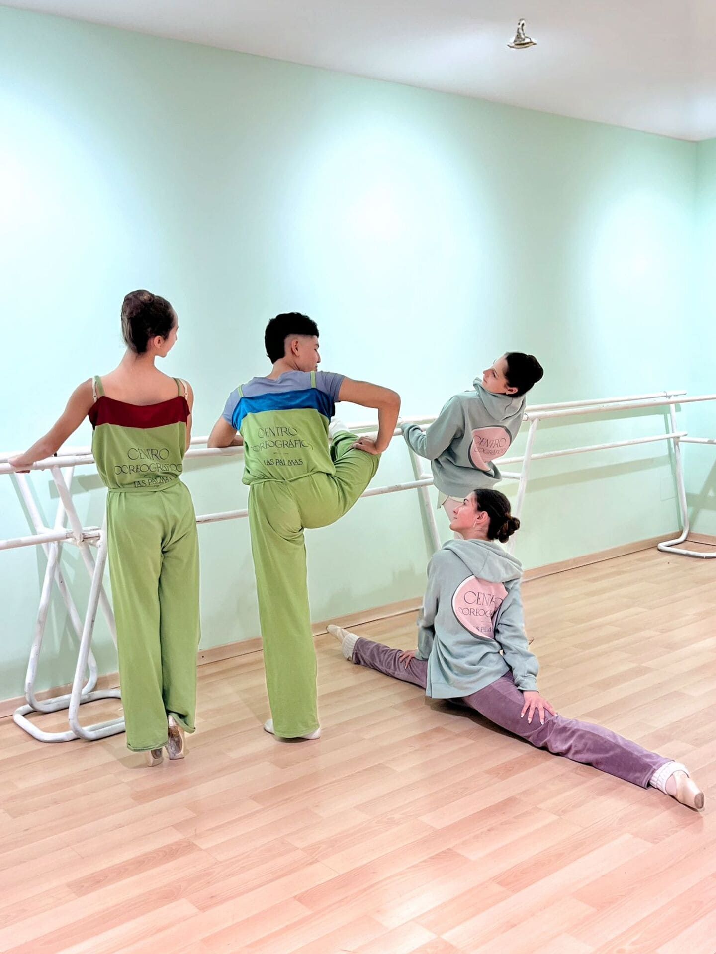 Tres bailarinas con pantalones verdes posan en las barras de ballet de un estudio; una de ellas está sentada en el suelo haciendo una apertura de piernas.