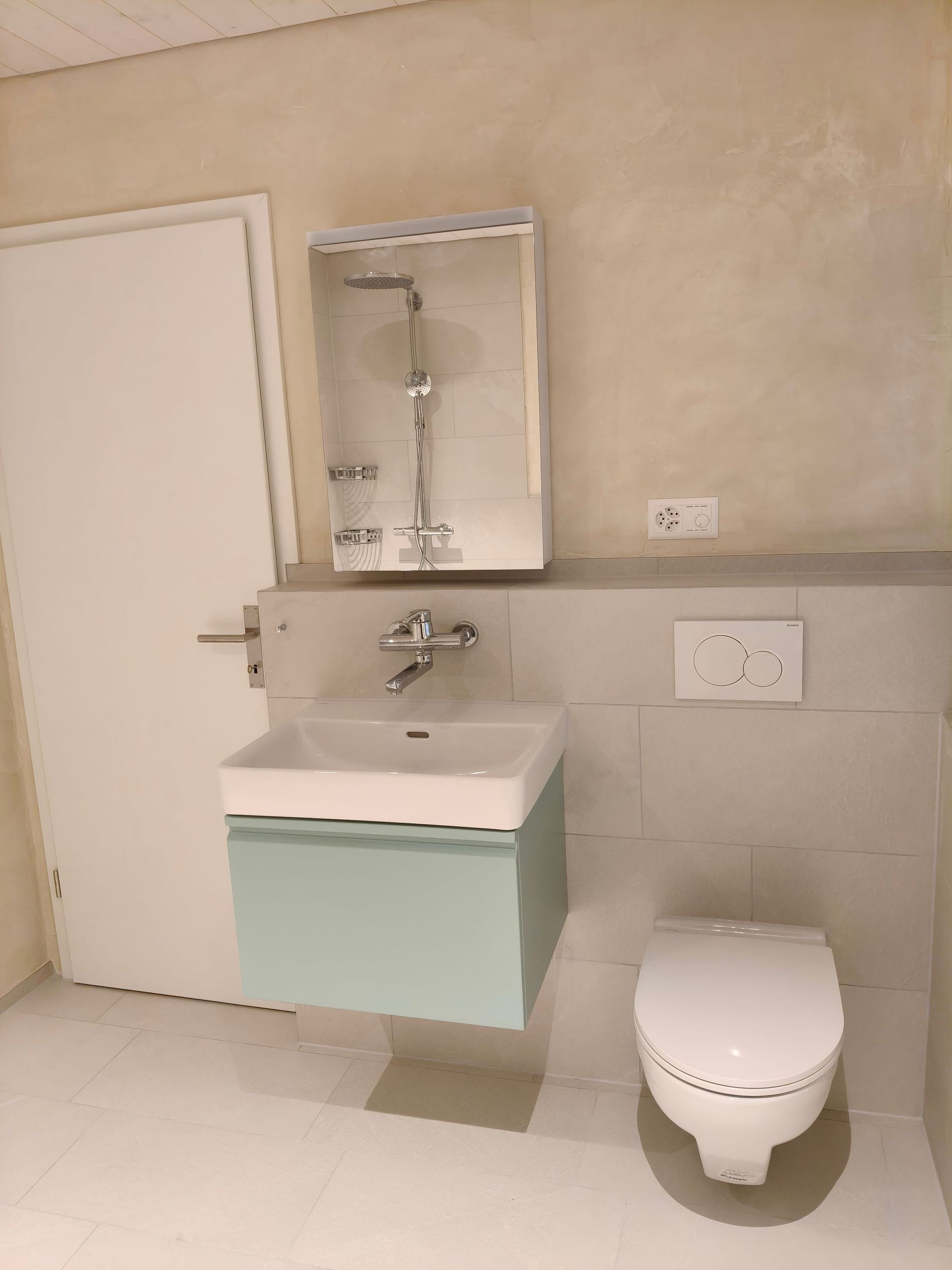 Ein modernes Badezimmer mit hellblauem Waschtisch, Toilette und Duschkopf. Weiße Tür auf der linken Seite.