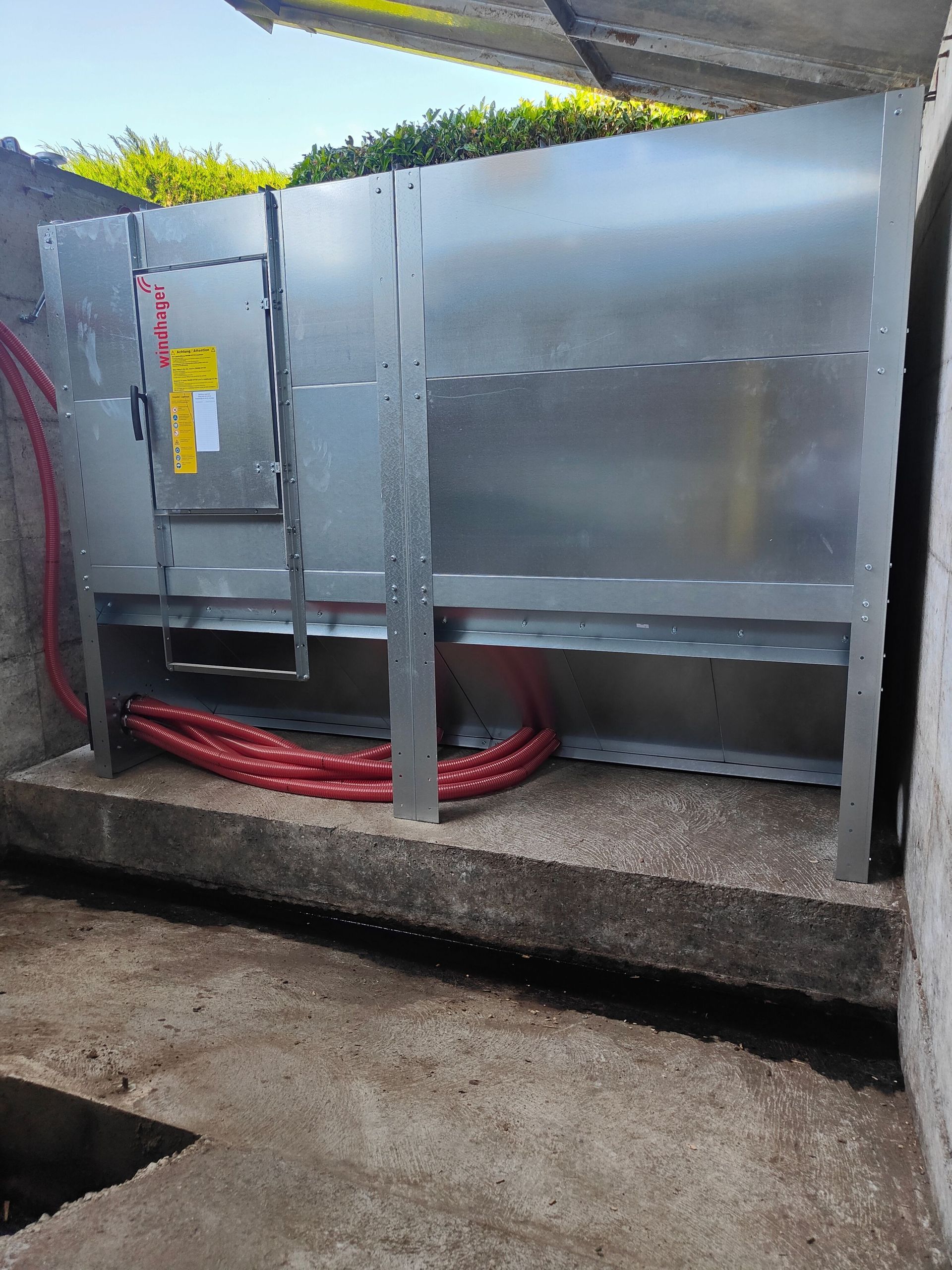 Metallische HVAC-Einheit an einer Wand, rote Rohre entlang des Betonsockels.