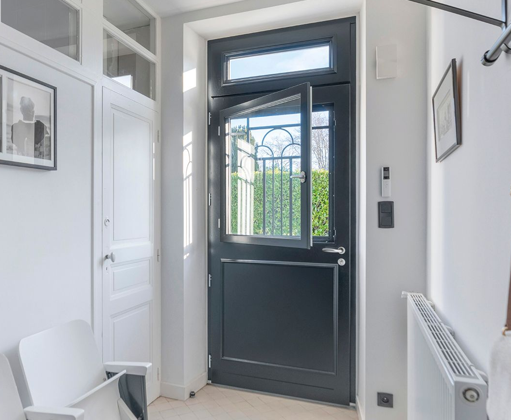 Porte hollandaise grise ouverte, donnant sur un portail extérieur. Intérieur blanc avec chaise et radiateur.