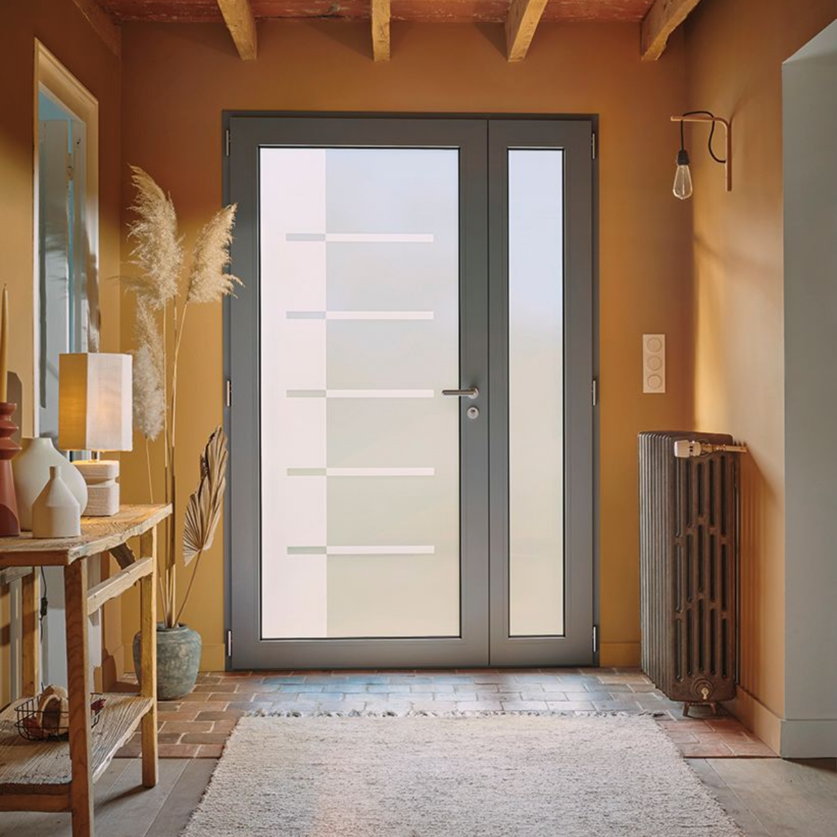 Entrée avec porte grise, fenêtre latérale, radiateur et table décorative. Les murs sont d'une chaude couleur orange.