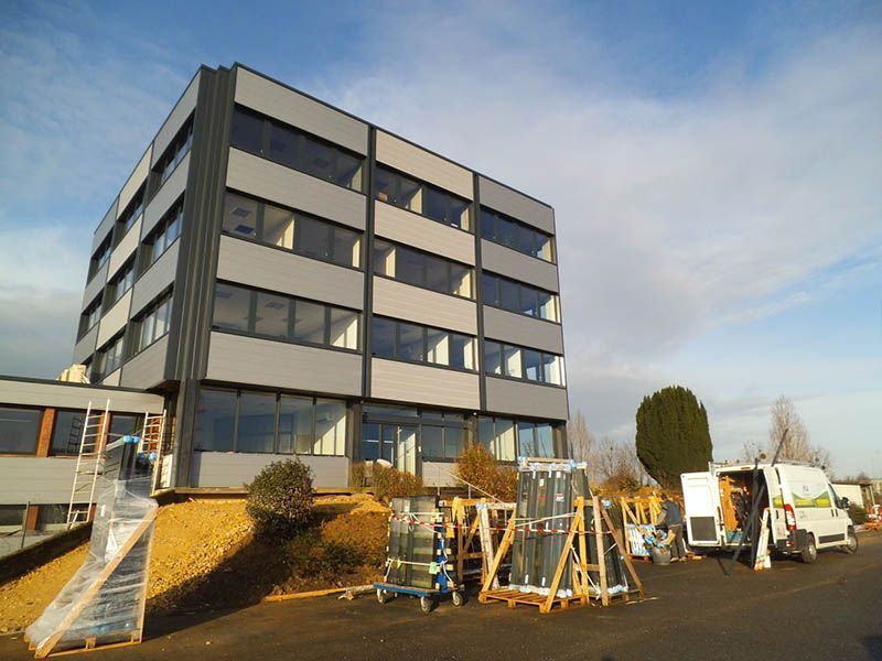 Immeuble de bureaux moderne gris ; des ouvriers déchargent de grandes vitres d'une camionnette par une journée ensoleillée.