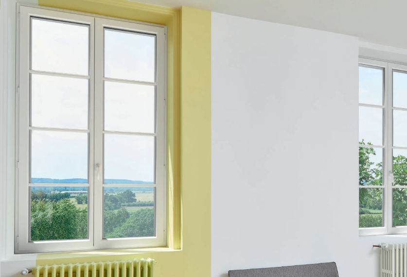Des fenêtres à cadre blanc dans une pièce aux murs blancs et jaunes, donnant sur un paysage verdoyant.