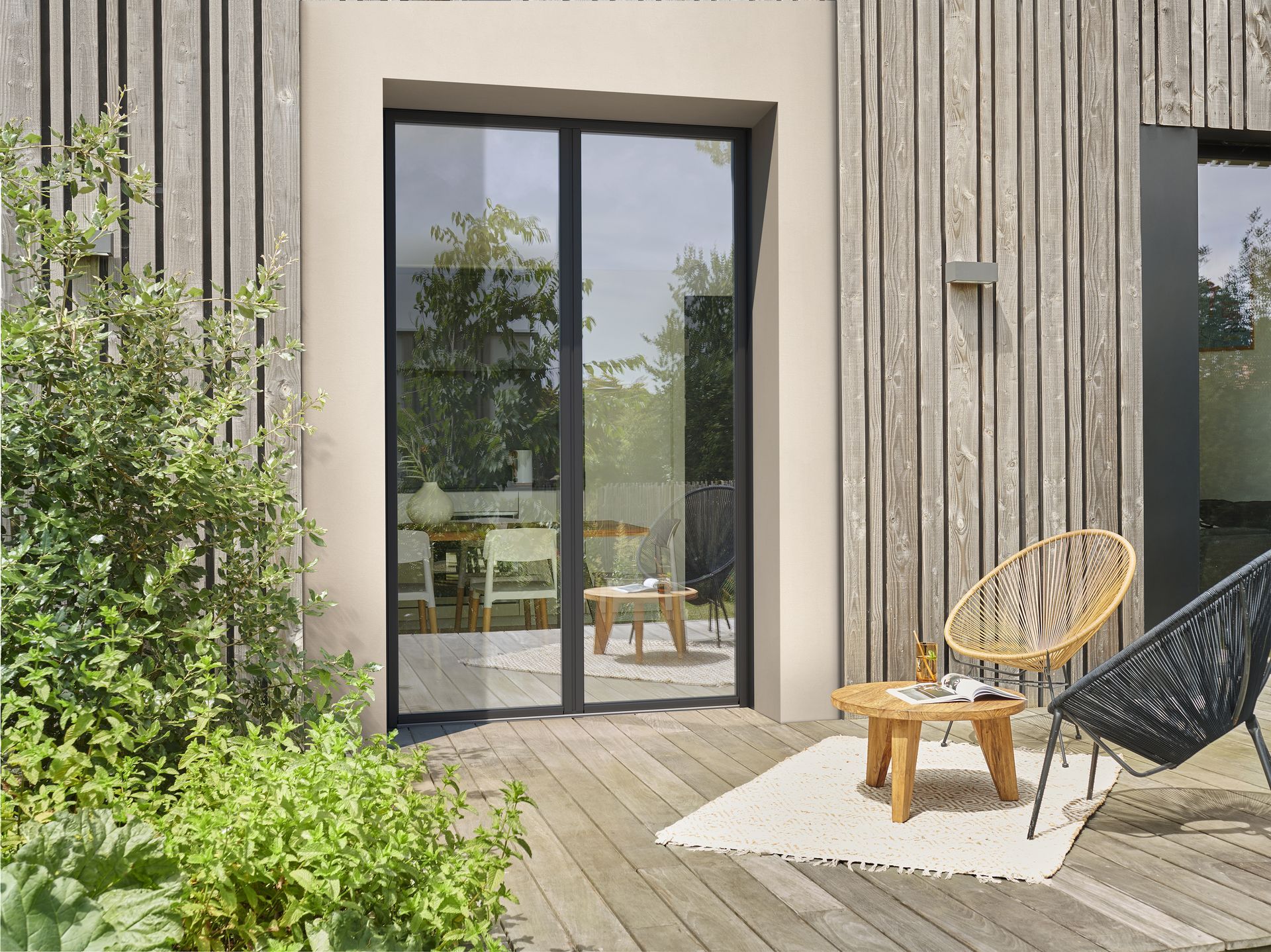 Porte-fenêtre coulissante donnant sur une terrasse en bois. Deux chaises, une petite table et des plantes sont présentes. Vue extérieure d'une maison.