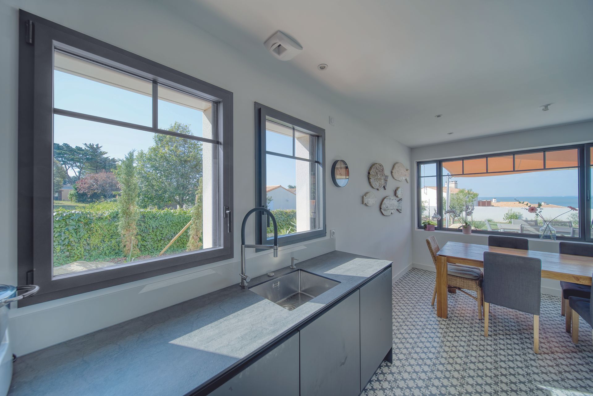 Cuisine avec placards gris foncé, évier et fenêtres donnant sur un jardin verdoyant et l'océan.