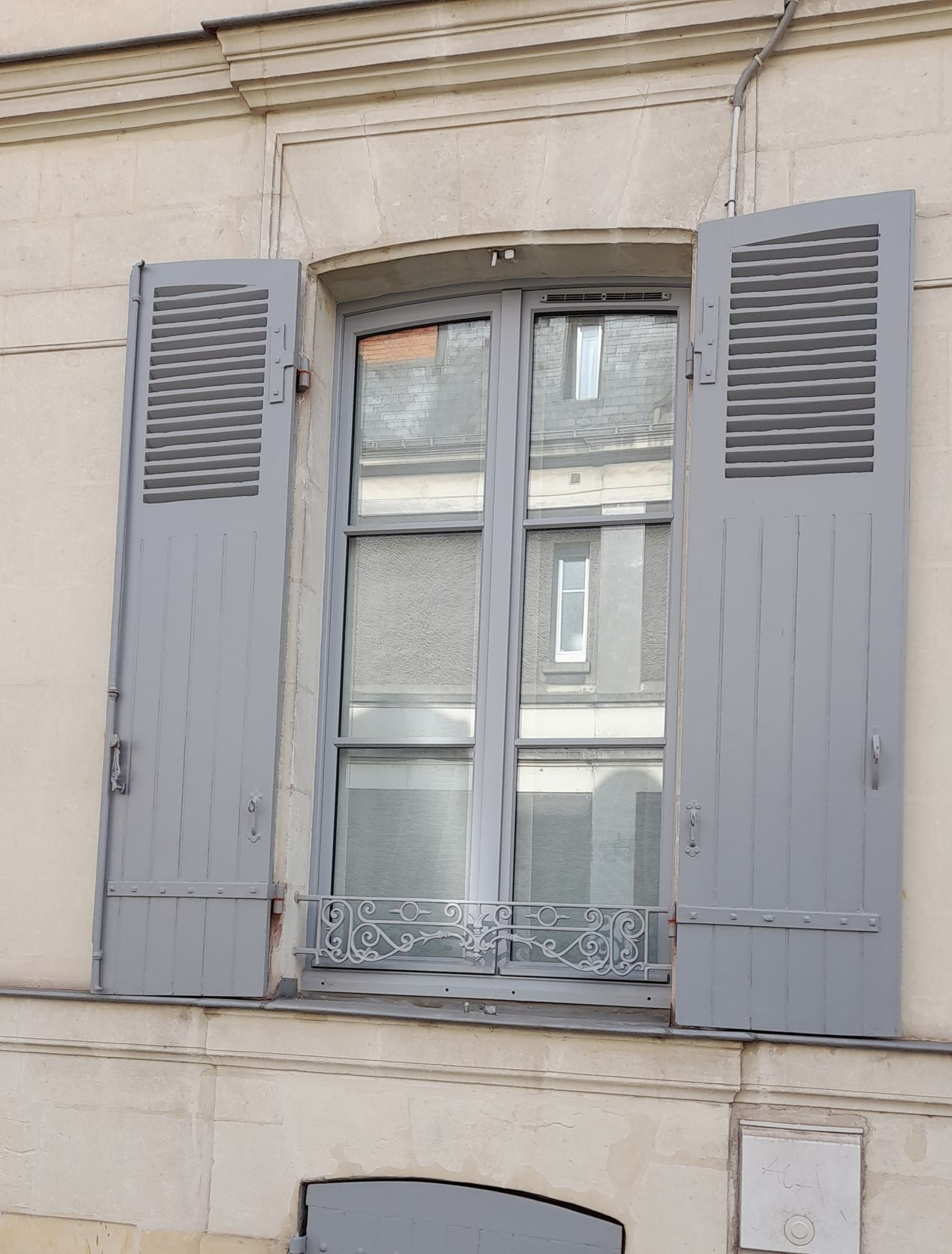 Fenêtre aux volets gris ouverts reflétant les bâtiments de l'autre côté de la rue.