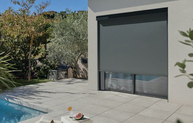 Un volet roulant gris foncé recouvre partiellement une fenêtre d'un bâtiment blanc, donnant sur une terrasse et une piscine.