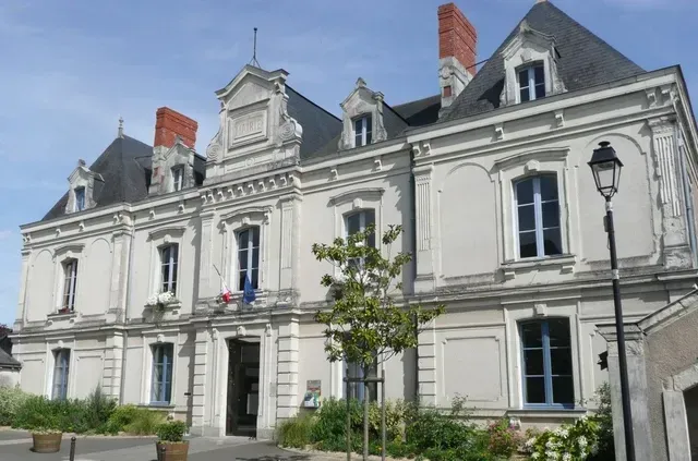 Bâtiment de deux étages de couleur crème, avec un toit foncé et des fenêtres à encadrement bleu ; drapeau français visible.