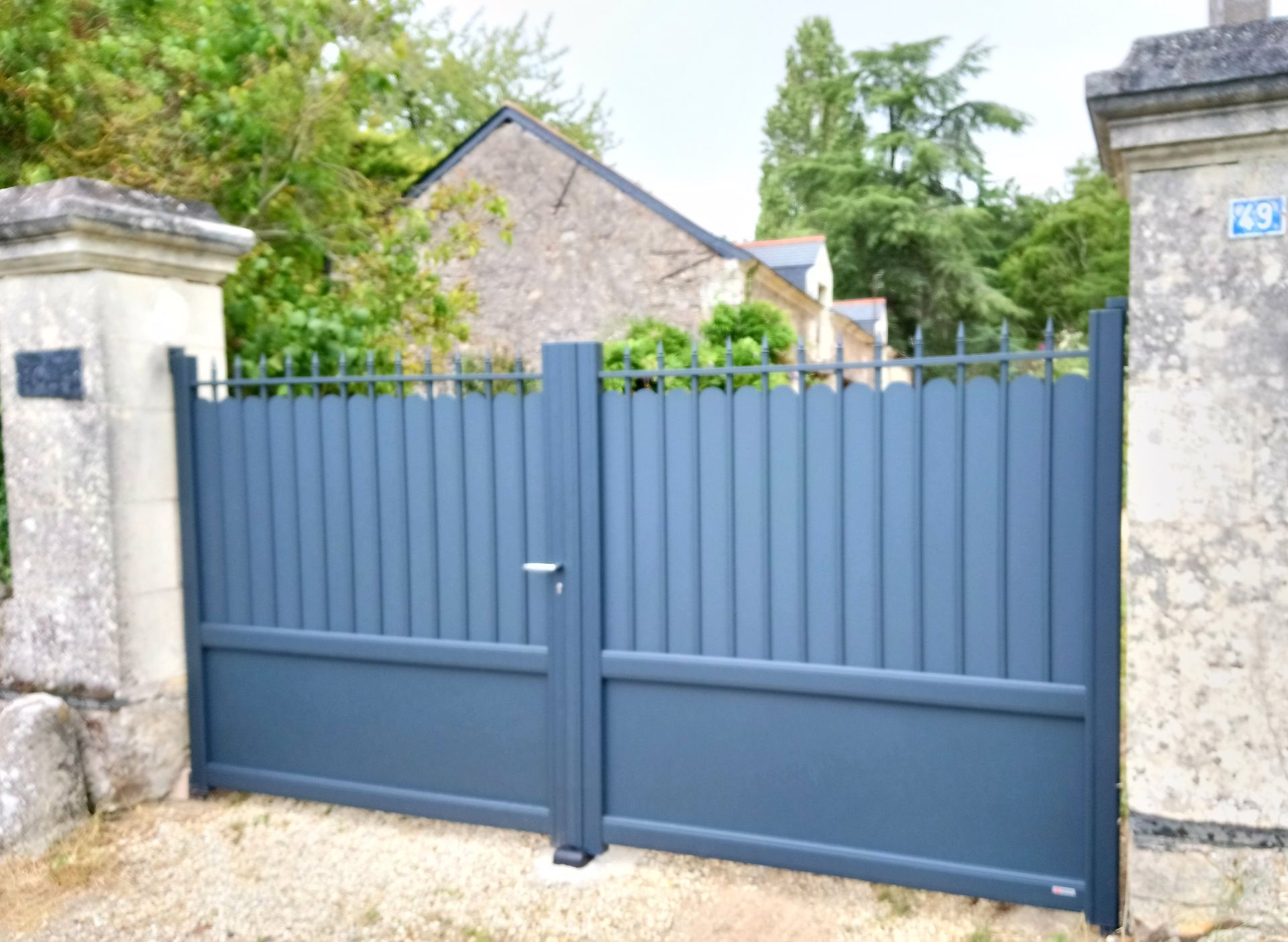 Portail en métal gris à barreaux verticaux, entre des piliers en pierre, menant à une maison.