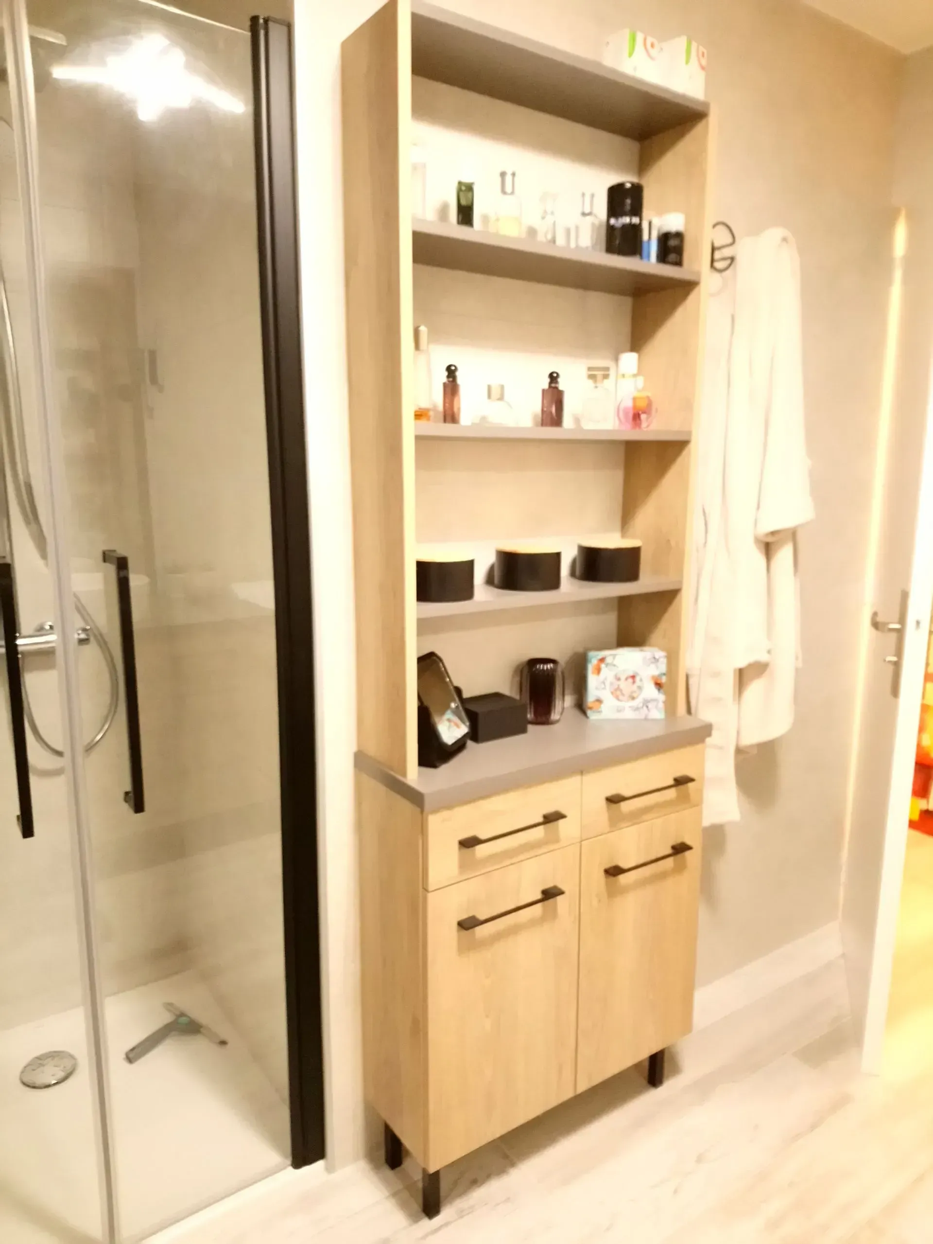 Meuble de salle de bain en bois, haut, avec étagères ouvertes, portes fermées et un peignoir accroché au mur.