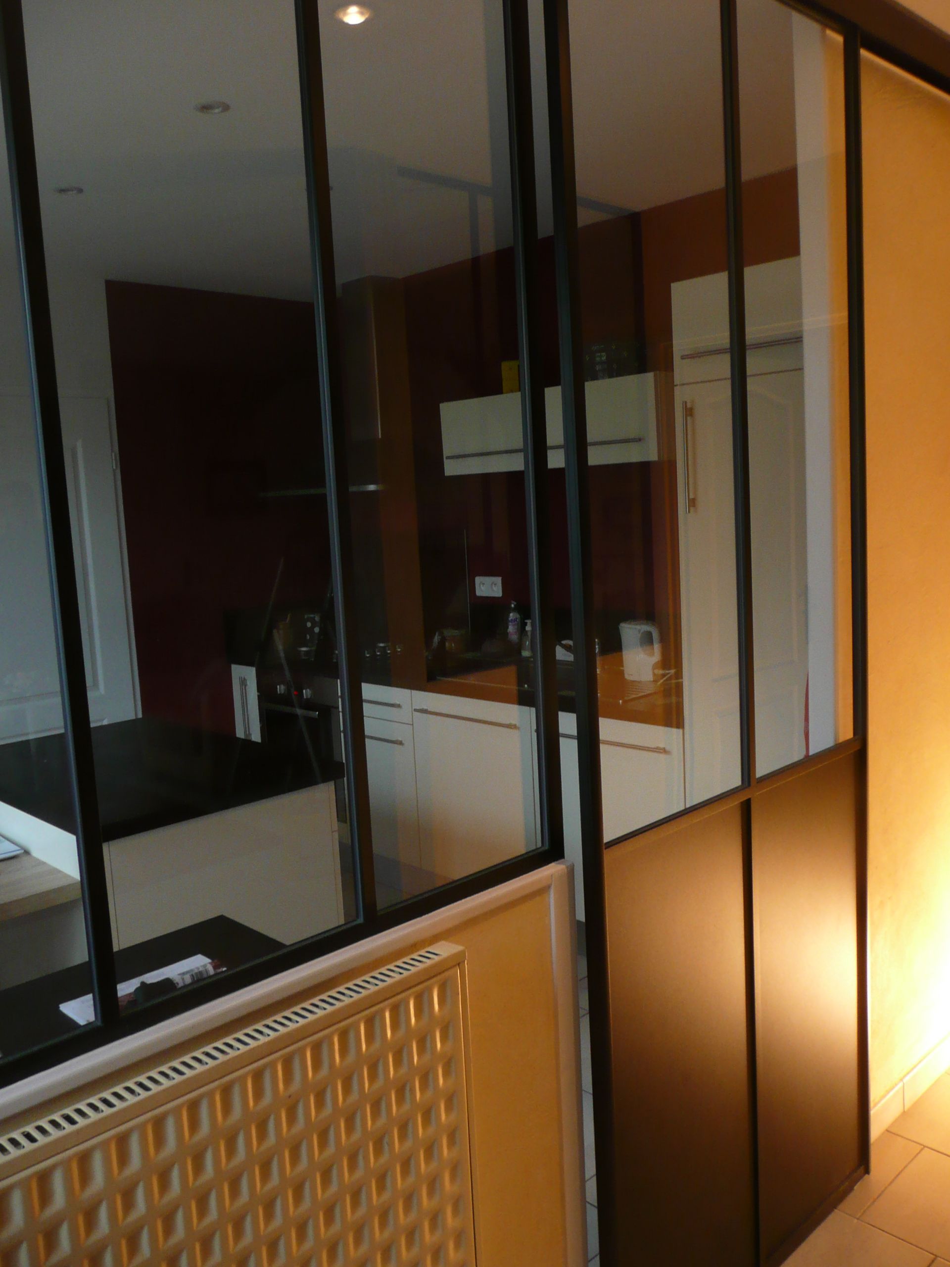 Une paroi vitrée à cadre noir sépare une cuisine aux placards blancs d'un coin salon équipé d'un radiateur.