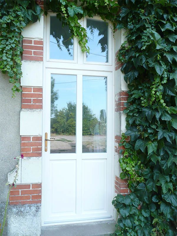 Porte blanche à panneaux de verre encadrée de lierre, de briques et de pierres, reflétant les arbres.