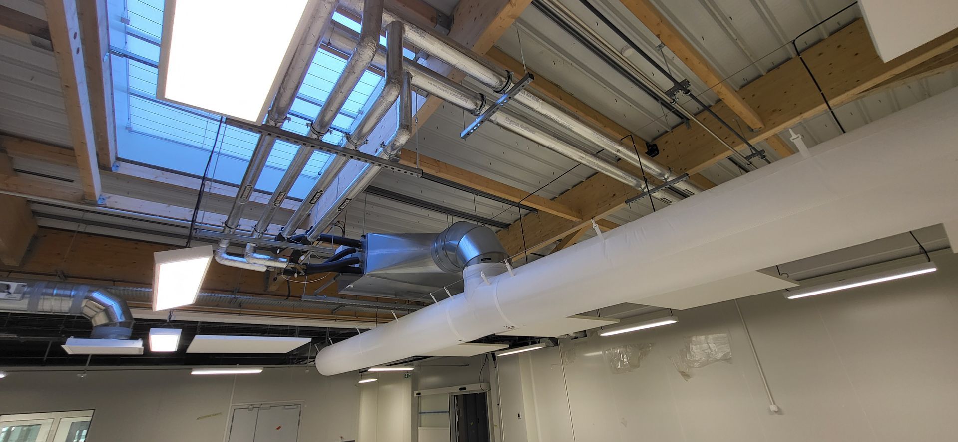 Pièce industrielle vide avec poutres apparentes au plafond, puits de lumière, conduits et murs blancs