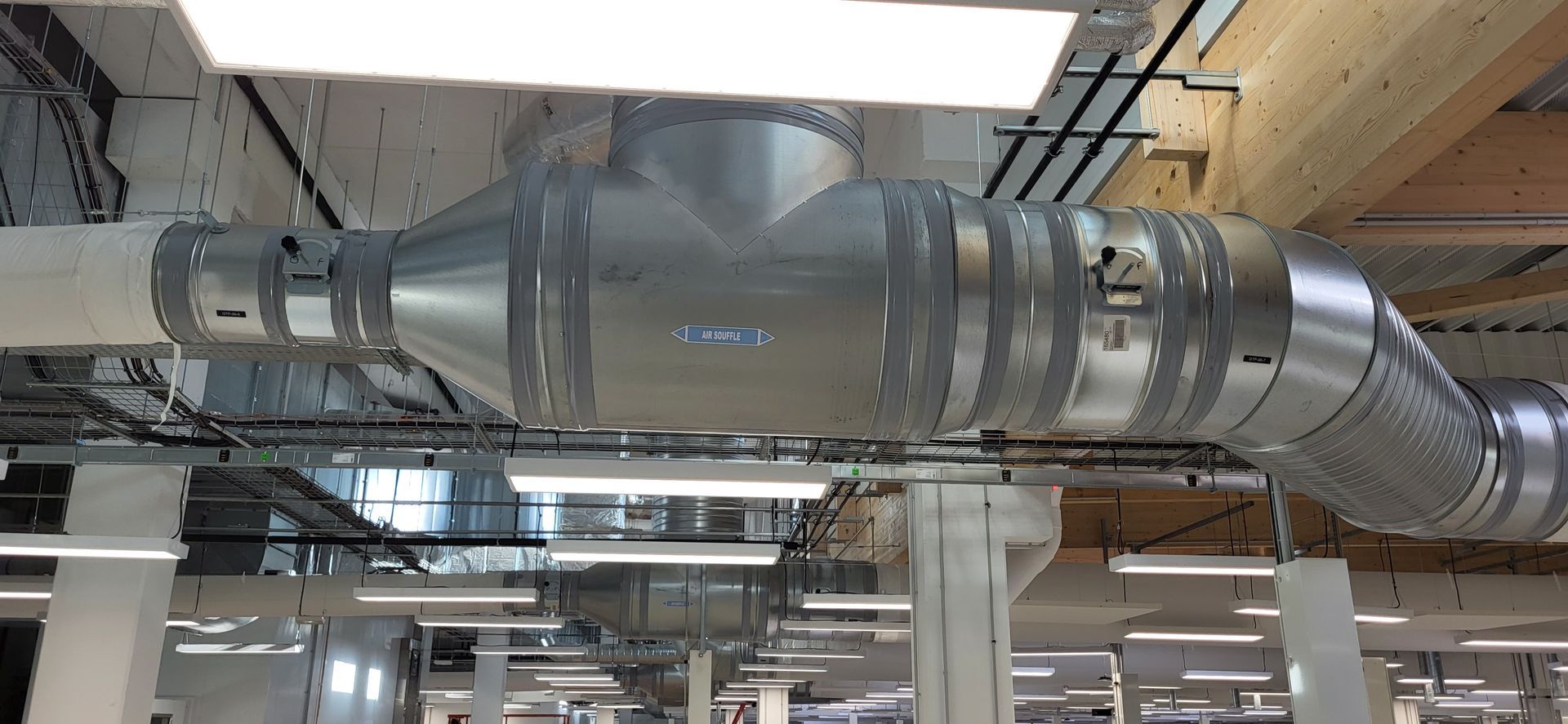 Conduits et tuyaux industriels à l'intérieur du plafond d'une usine, avec une grande unité de ventilation grise visible.
