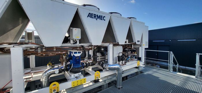 De grandes unités de climatisation AERMEC blanches installées sur un toit, avec des tuyauteries bleues et des équipements industriels au premier plan.
