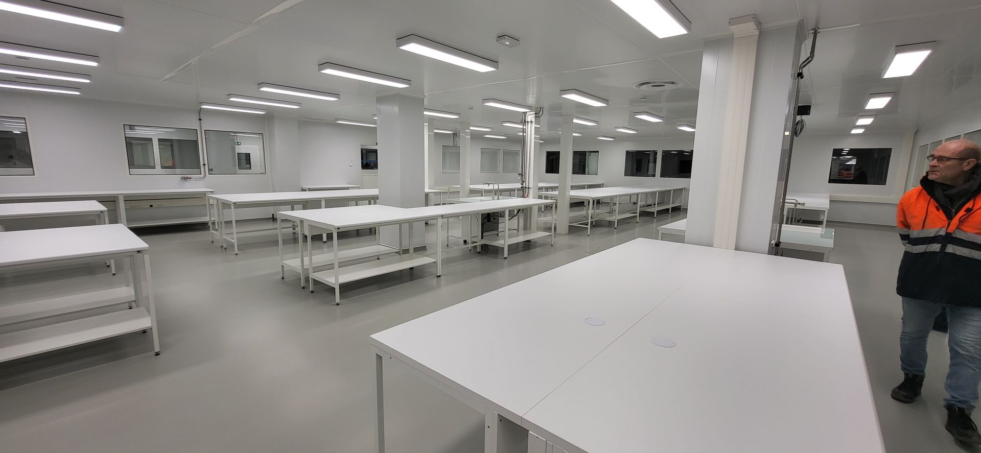 Laboratoire ou salle d'usine lumineuse avec des tables et des étagères blanches, et une personne en tenue de sécurité orange debout à droite
