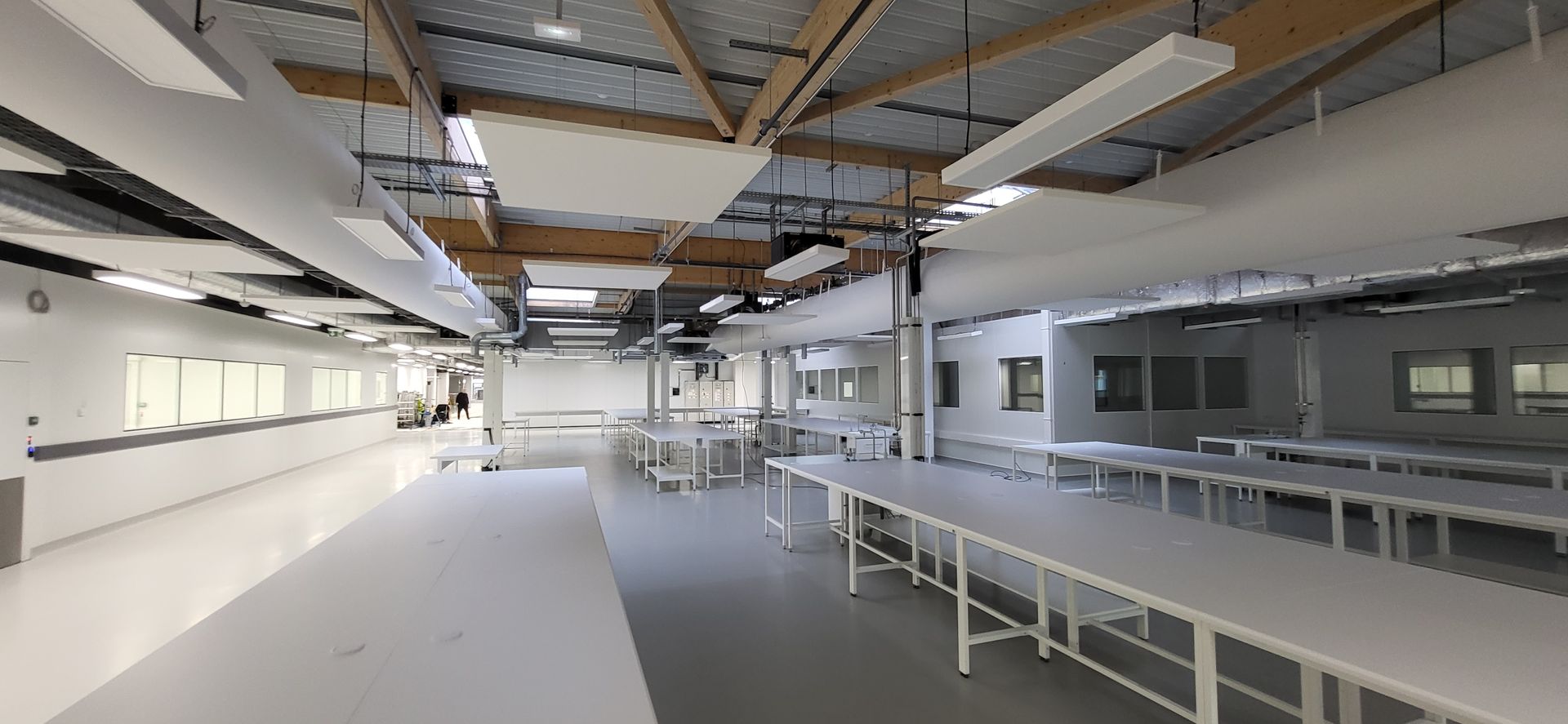 Espace de travail intérieur lumineux et vide, avec de longues tables, des murs blancs et des poutres apparentes au plafond.