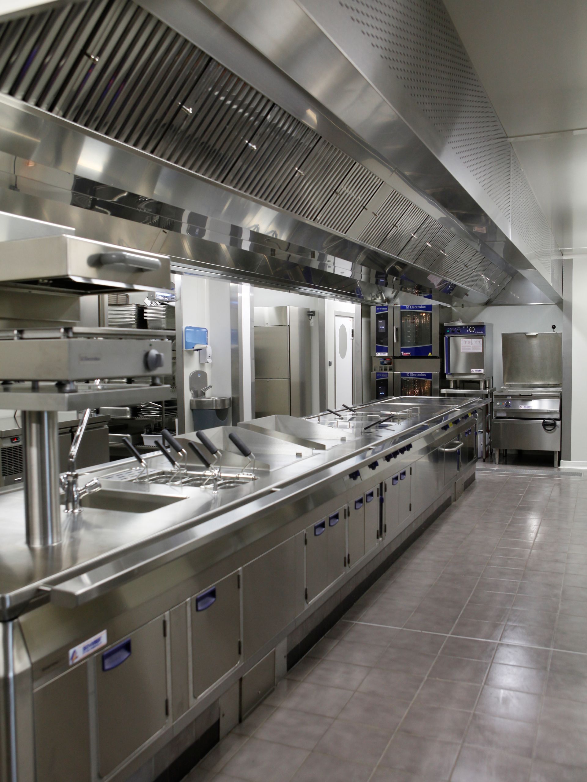 Cuisine commerciale en acier inoxydable avec longs plans de travail, éviers, fours et hottes d'extraction métalliques.
