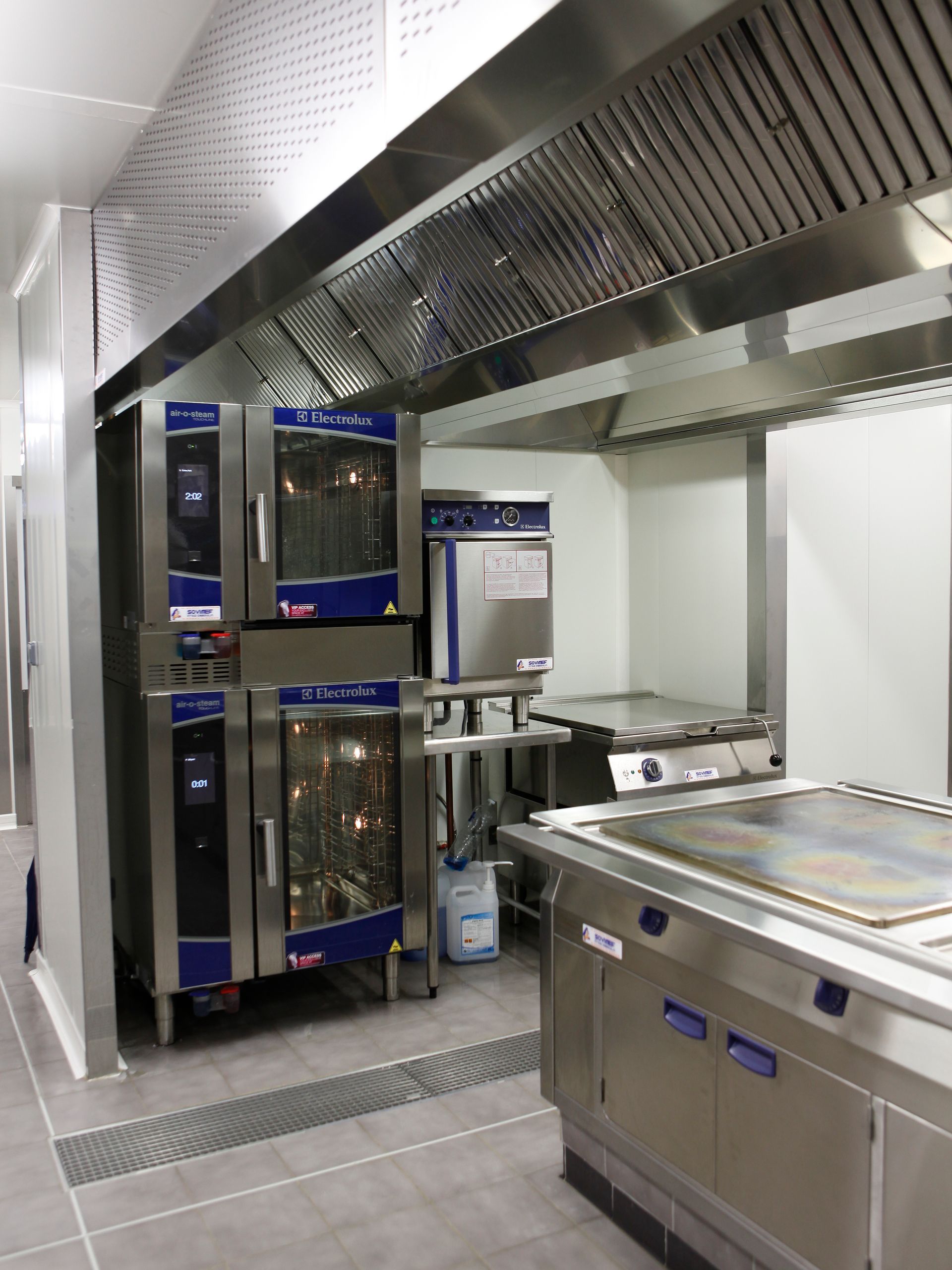 Cuisine professionnelle équipée d'appareils de cuisson en acier inoxydable et de fours à bordure bleue sous une hotte aspirante.