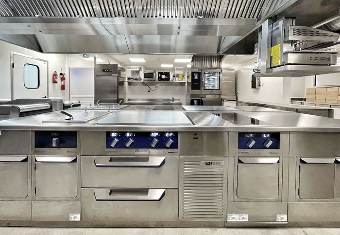 Cuisine professionnelle en acier inoxydable avec fours, éviers et comptoirs, éclairée par un éclairage zénithal puissant.