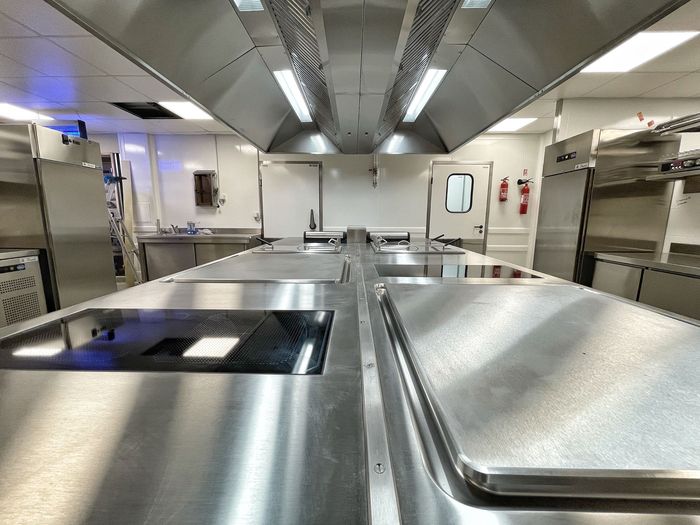 Cuisine professionnelle avec comptoirs en acier inoxydable, fours et hottes aspirantes, éclairée par une lumière vive.
