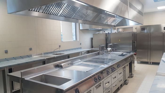 Cuisine professionnelle avec comptoirs en acier inoxydable, fours, éviers et grande hotte aspirante.