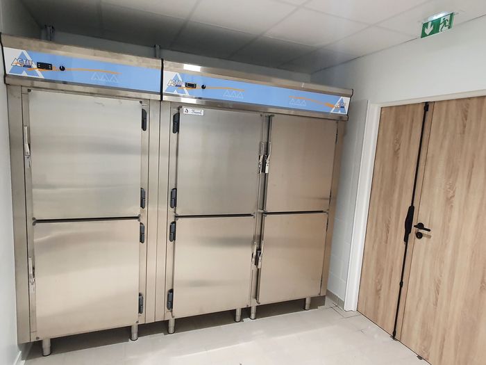 Toilettes publiques avec portes de cabine en acier inoxydable, murs carrelés blancs et une porte en bois sous un panneau de sortie