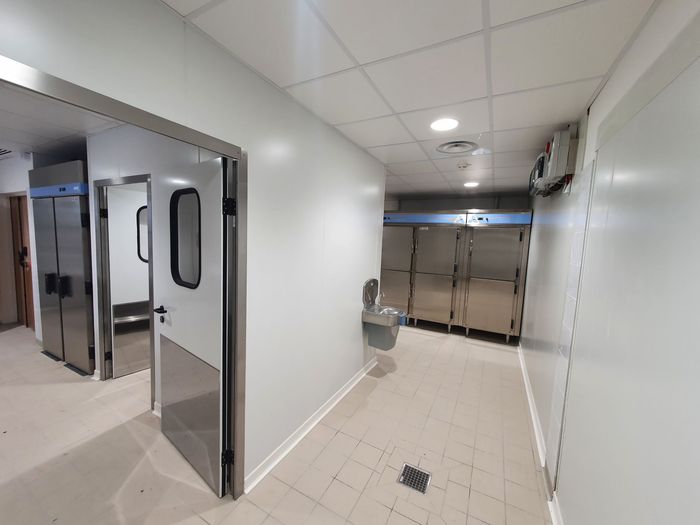 Couloir d'hôpital vide, portes ouvertes, murs blancs, sol beige et un lavabo au fond.