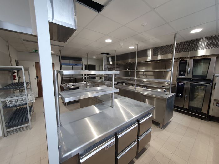 Cuisine commerciale lumineuse en acier inoxydable avec comptoirs de préparation, étagères, fours et sol carrelé.