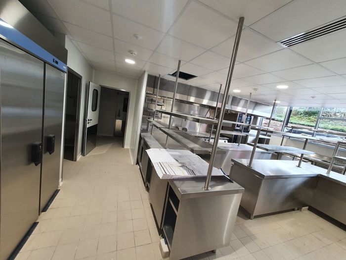 Cuisine commerciale vide avec comptoirs, étagères et réfrigérateurs en acier inoxydable, éclairée par un plafond lumineux.