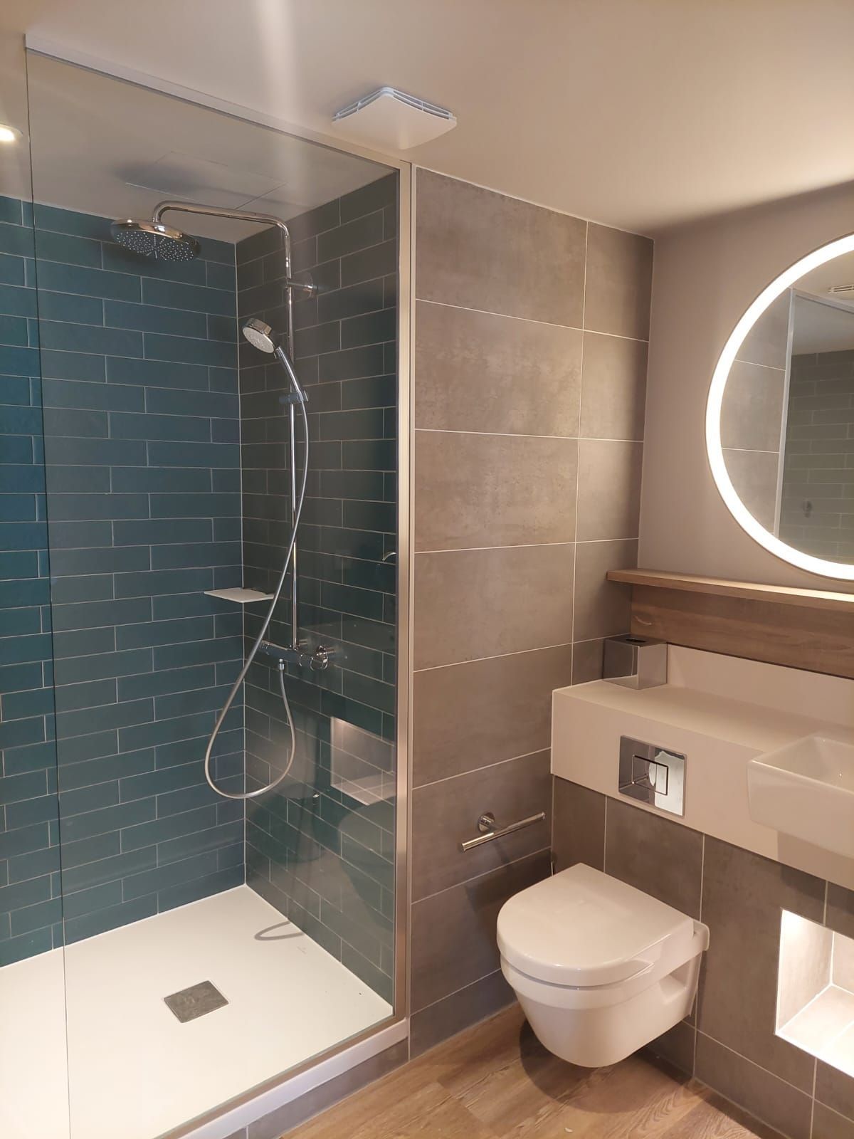 Salle de bains moderne avec douche en verre, carrelage foncé, toilettes, meuble-lavabo et miroir rond éclairé