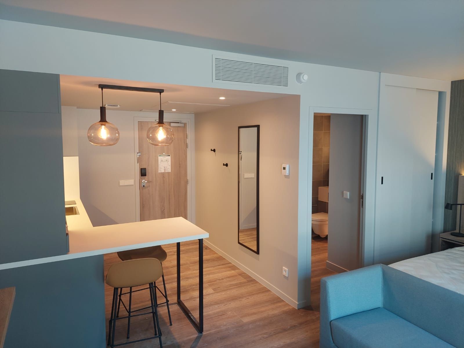 Studio moderne avec kitchenette, tabourets de bar, suspensions lumineuses et canapé bleu. Vue