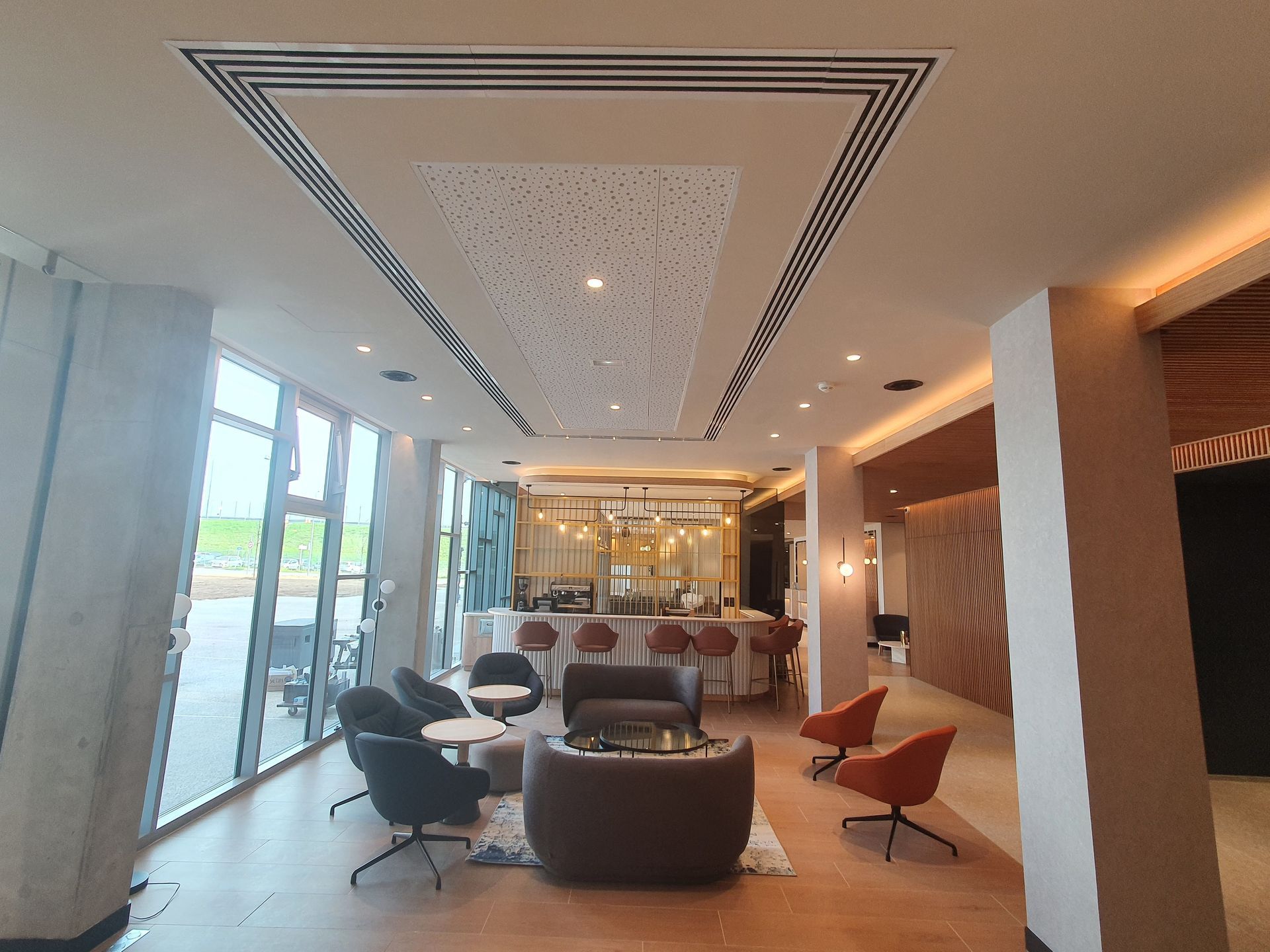 Salon d'hôtel moderne avec fauteuils, tables et un plafond lumineux grâce à des baies vitrées allant du sol au plafond.