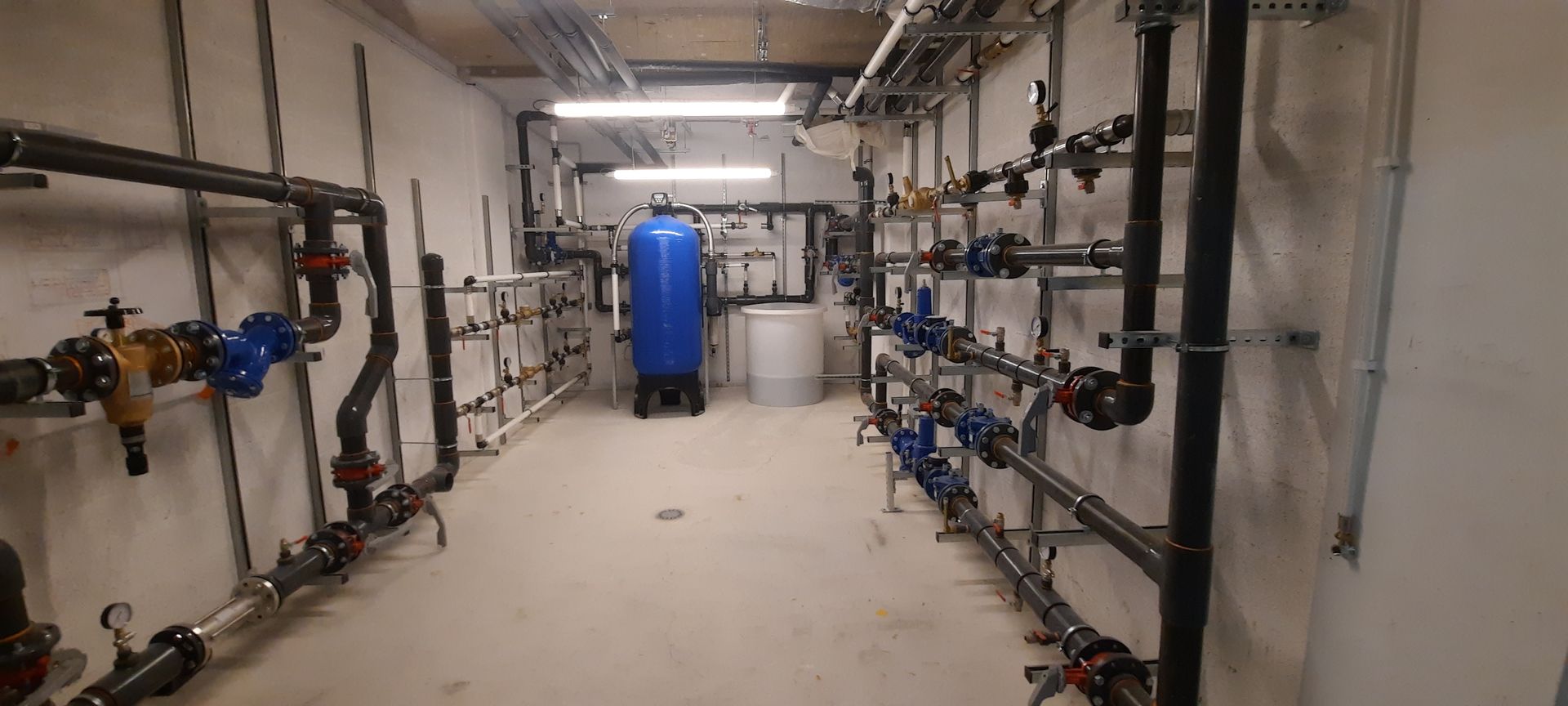 Local technique industriel avec des tuyaux et des vannes le long des deux murs et un réservoir bleu au fond.