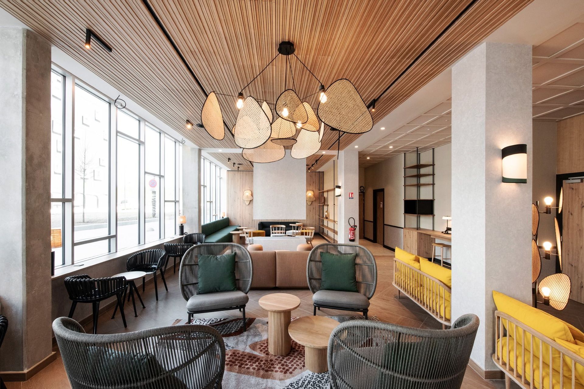 Salon moderne avec luminaires suspendus en osier, chaises vertes, canapé jaune et grandes fenêtres