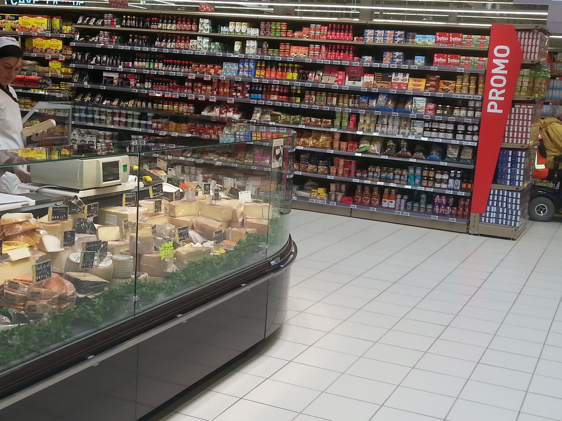 Allée d'un supermarché avec des étagères de produits emballés et un comptoir de charcuterie au premier plan