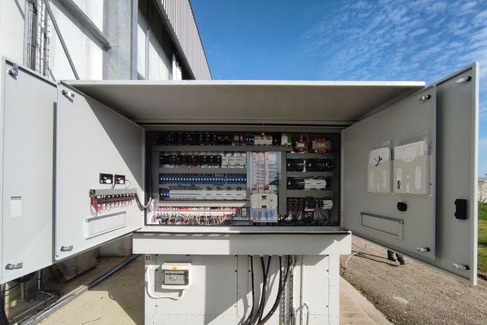Armoire électrique ouverte avec câblage et composants à l'extérieur, contre un mur du bâtiment.
