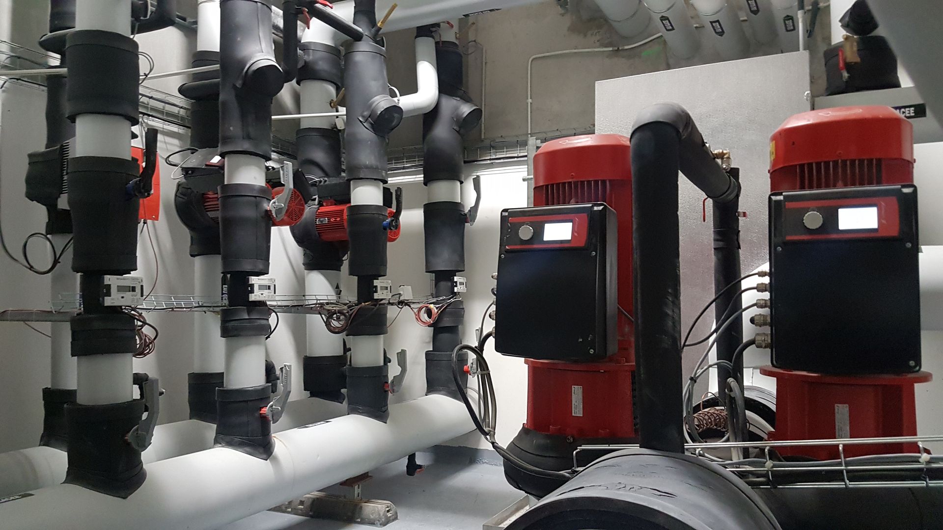 Local technique industriel avec pompes rouges, tuyaux argentés et équipements isolés fixés au mur