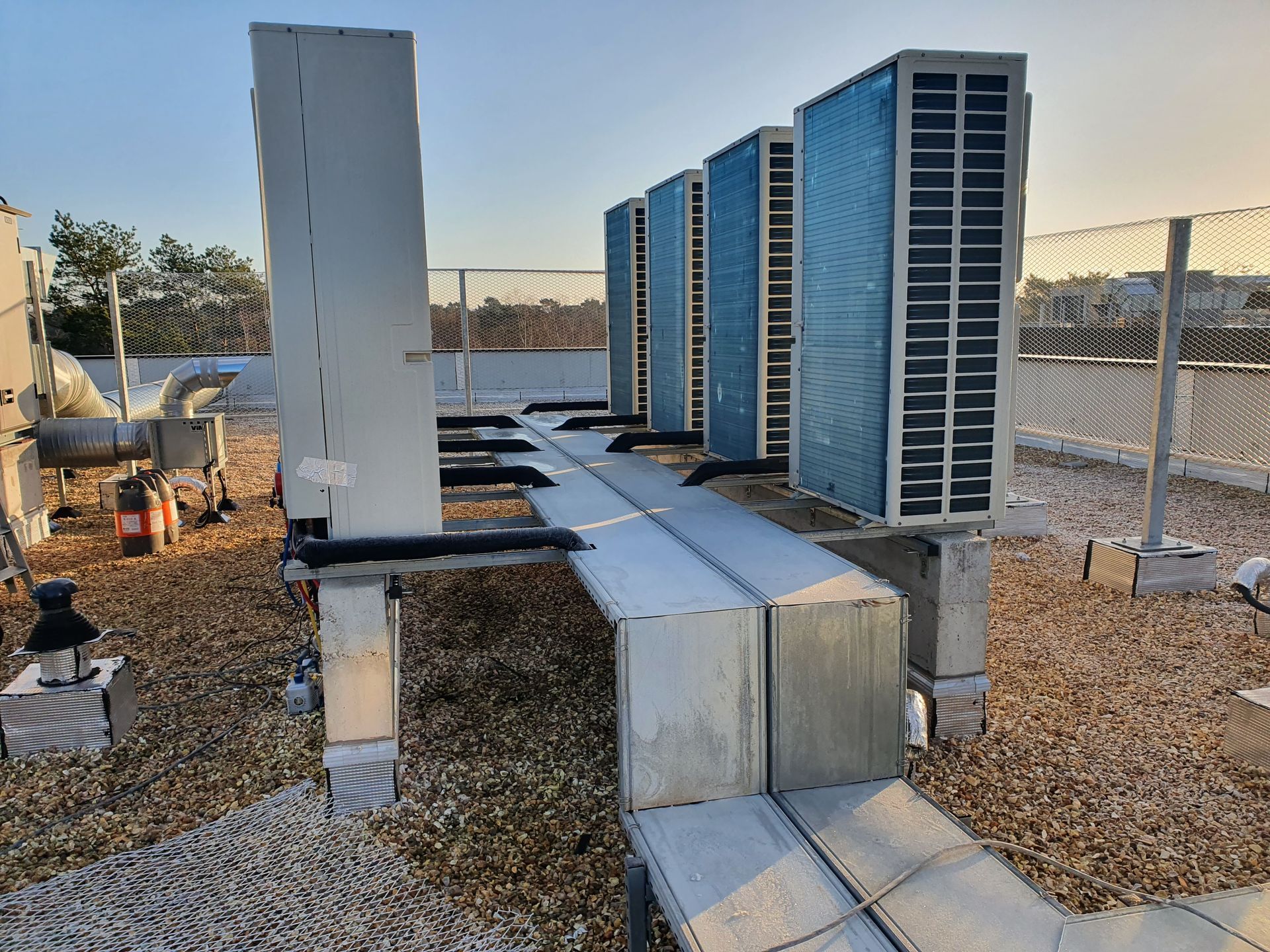 Unités de climatisation extérieures et conduits d'aération sur un toit en gravier, à côté d'une clôture en grillage.