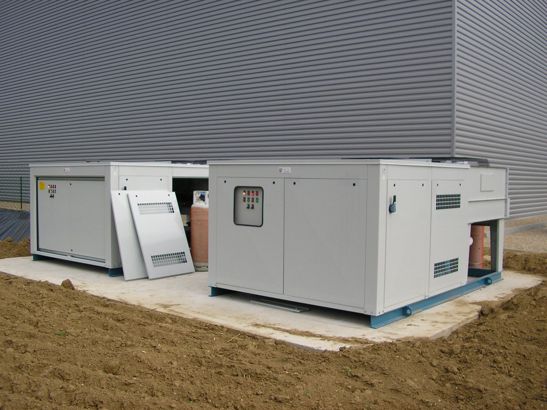 Unités d'équipement électrique industriel à côté d'un bâtiment, avec une dalle de béton et un premier plan en terre.