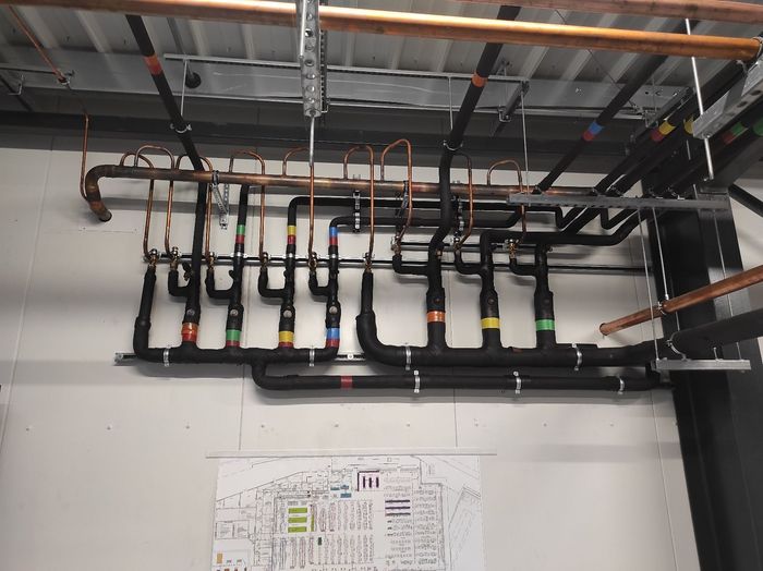 Plafond industriel avec rangées de tuyaux noirs, isolation colorée et conduits électriques dans une buanderie