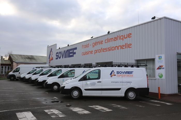 Une rangée de fourgonnettes blanches de service stationnées devant un bâtiment arborant le logo du SOVIMF et des panneaux de signalisation français orange.
