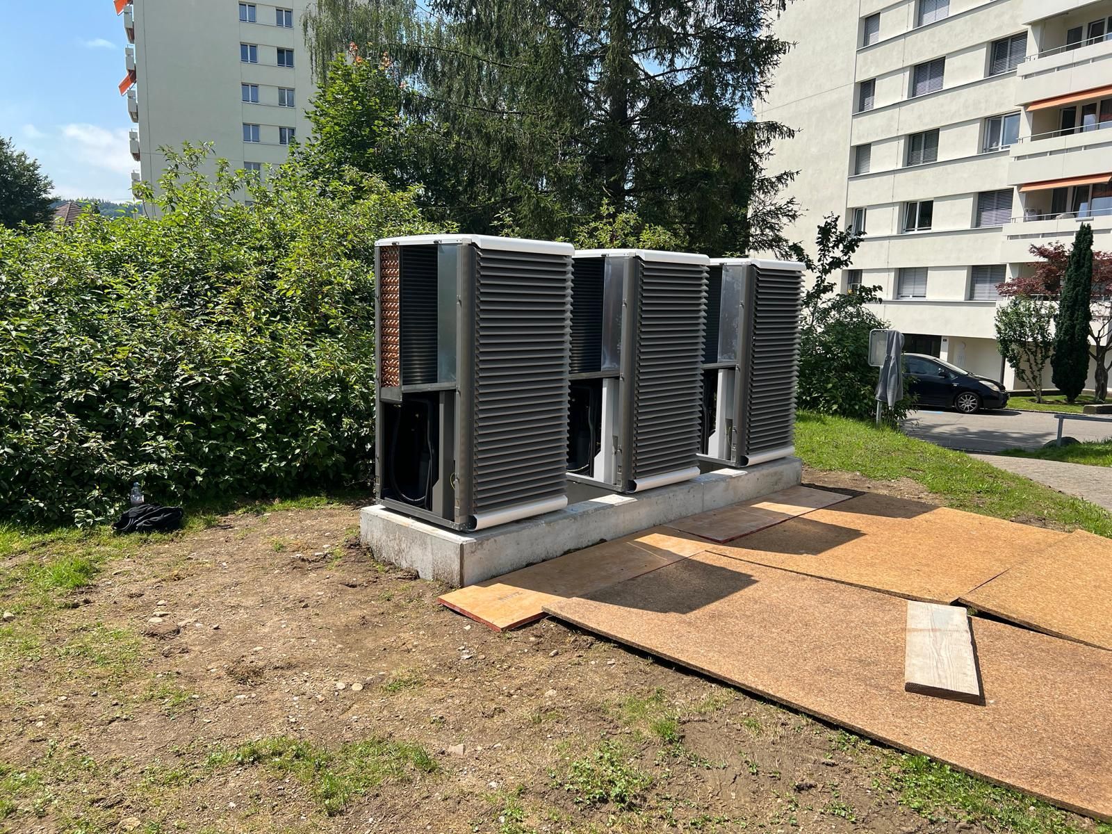 Raum mit schwarzem Warmwasserbereiter, weißem Tank und abgedecktem Gerät. Wände und Boden aus Beton.