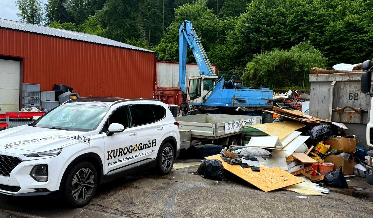Weißer Geländewagen mit Logo neben einem Müllhaufen, im Hintergrund ein blauer Bagger und ein rotes Gebäude.