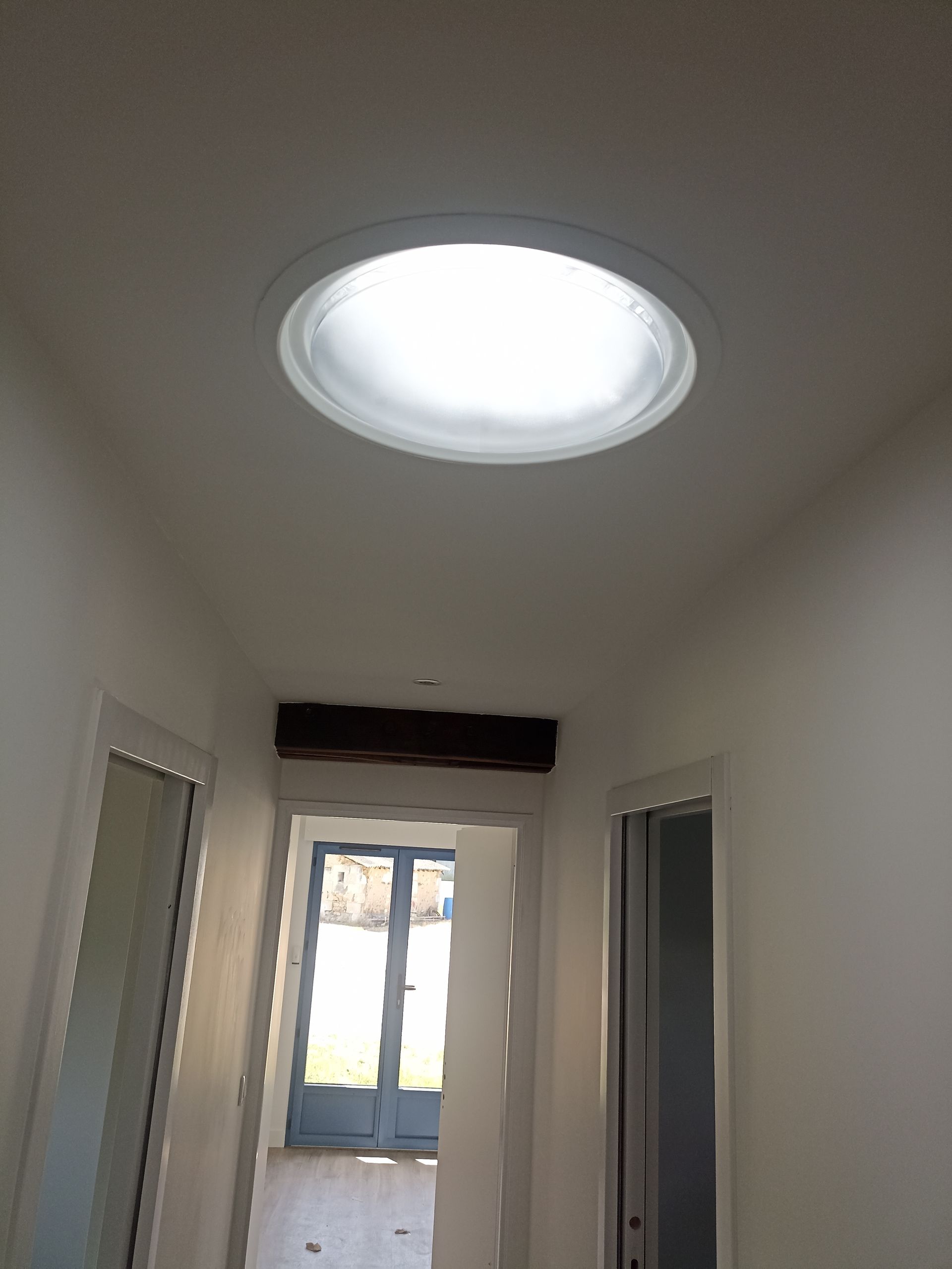 Couloir étroit aux murs blancs, plafond avec puits de lumière et porte lumineuse au fond.