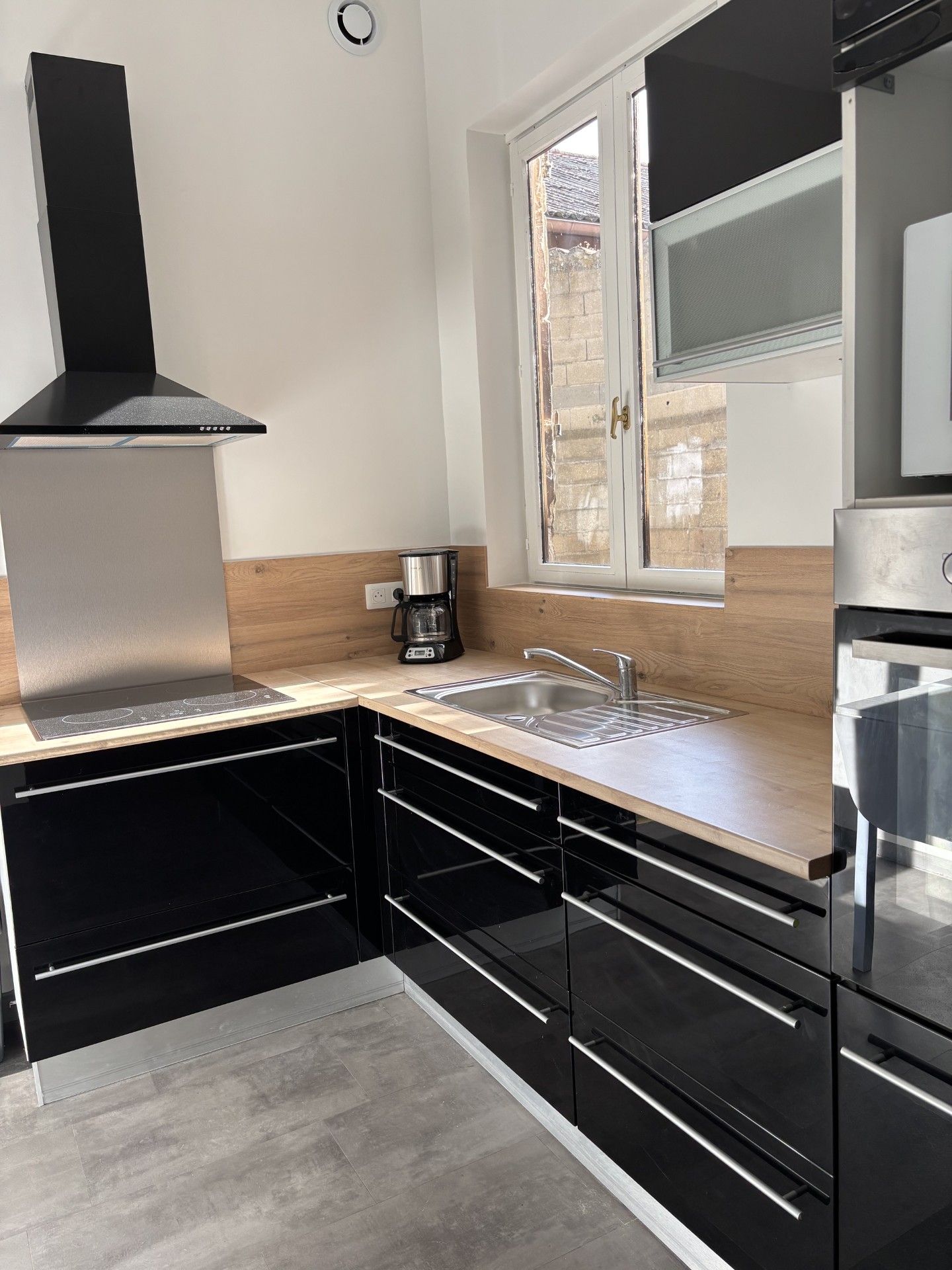 Cuisine moderne noire et inox avec comptoirs en L, fenêtre et hotte aspirante.