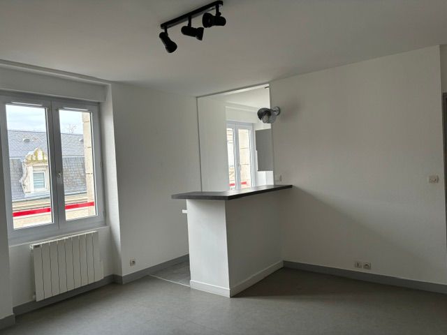 Pièce vide avec murs blancs, sol gris, fenêtres, radiateur et petit comptoir de bar.